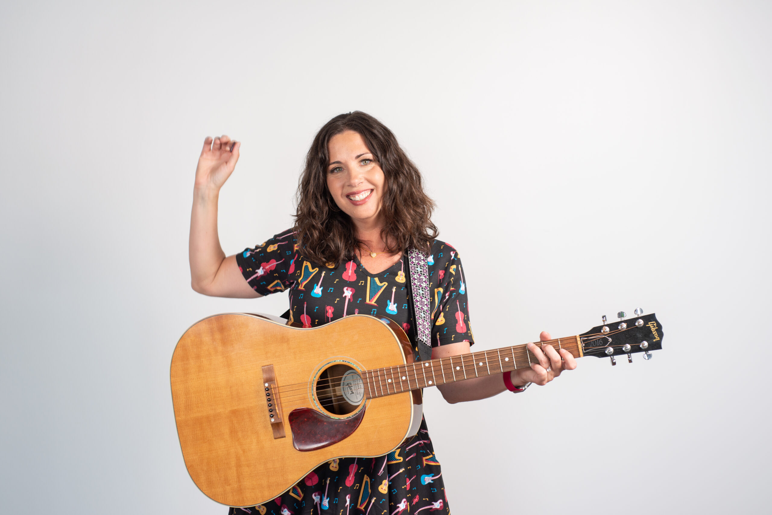 Stacey Peasley, a smiling white woman with brown hair, stands with a guitar. Her right hand is up like she just finished strumming, and she is wearing a dress patterned with different musical instruments